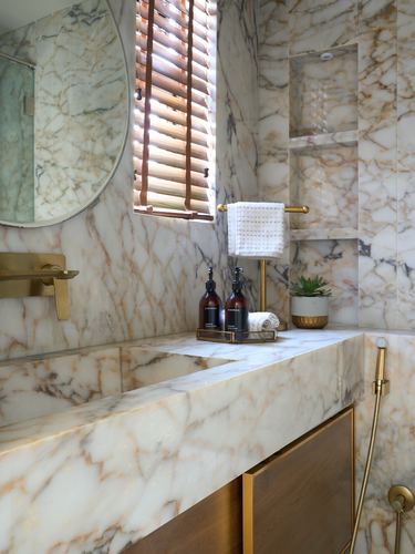 Marble bathroom vanity with brass fittings, round mirror, and wood-slat window blinds