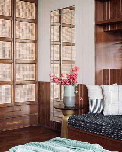 Corner of bedroom with cane wardrobe, mirror, vase of flowers, and ribbed wood daybed