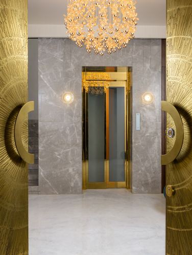 Textured brass doors open onto a marble foyer with a brass-framed elevator and chandelier.