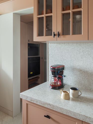Blush pink kitchen corner with stone counter, coffee maker, mug and creamer, glass cabinets above