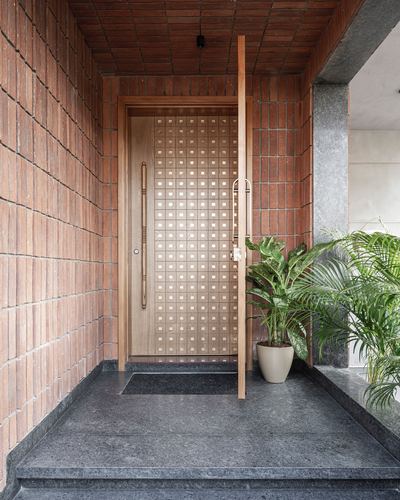 Wooden entry door with grid pattern set in brick-clad porch, granite flooring, potted palms beside it