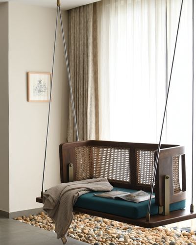 Wooden cane-paneled hanging swing with teal cushion over pebble flooring near sheer curtains