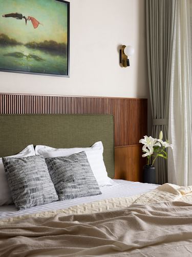 Bed with olive headboard against fluted wood panelling, sconce, painting, and vase of lilies beside curtained window