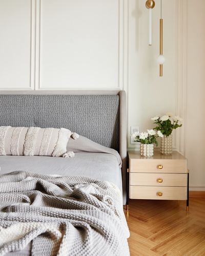 Grey upholstered bed beside a light wood nightstand with flowers and brass pendant lights
