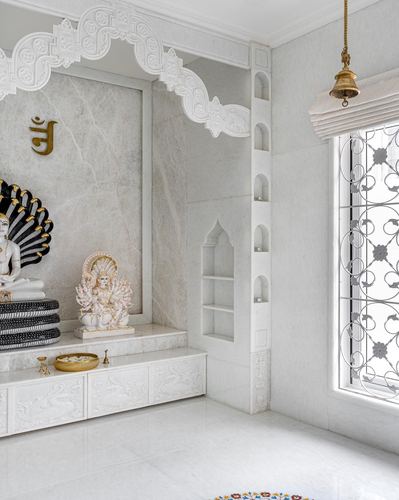 White marble pooja room with carved arch, deity idols, and a jali window with hanging brass bell