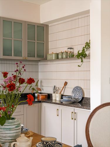 Kitchen corner with green cabinetry, vertical tiled backsplash, open shelf and floral bouquet