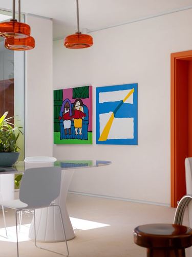 Dining nook with glass table, white chairs, two pop-art paintings and amber pendant lights beside orange doorway