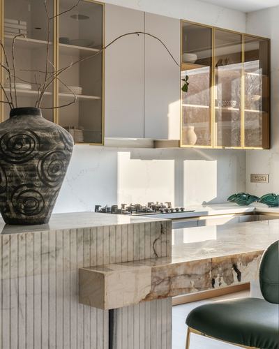 Kitchen counter with fluted stone island, gold-trimmed glass cabinets, large ceramic vase and dried branches