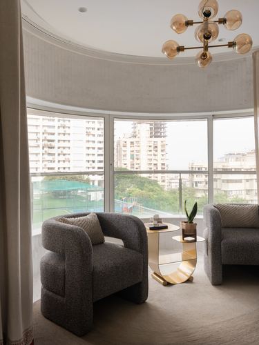 Two grey boucle armchairs face a curved window with brass side table and city view