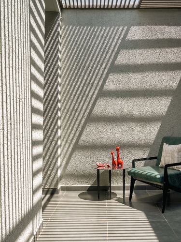 Sunlit balcony corner with shadow stripes on textured wall, green armchair and red figurines on side table