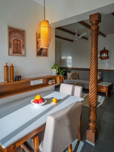 Dining table beside a carved wooden column with cane pendant light and framed Rajasthani door art