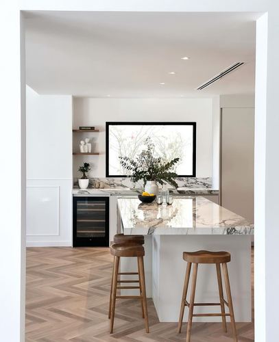 White kitchen island with marble waterfall countertop, wooden stools, and herringbone floor seen through doorway
