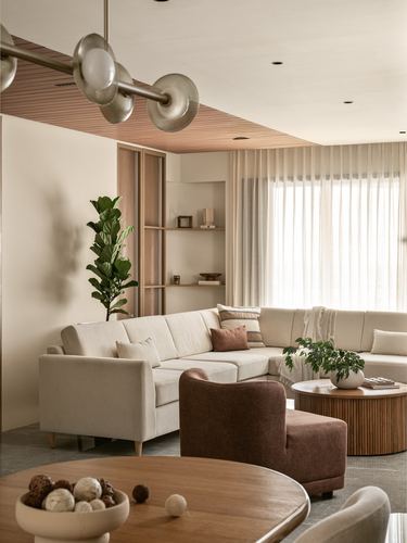 Beige sectional sofa, brown armchair, and round wood coffee table in a softly lit living room with sheer curtains.