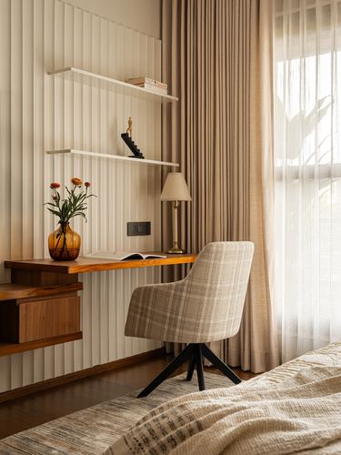 Fluted white wall with floating wood desk, plaid chair, and sheer curtained window in a bedroom study nook