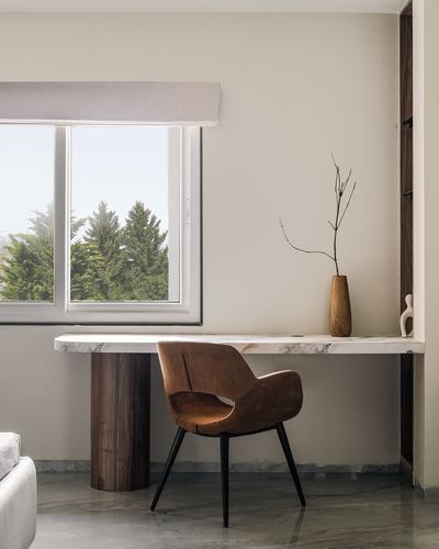 Marble desk on a cylindrical wood leg beneath a window, with a tan chair and wood vase.