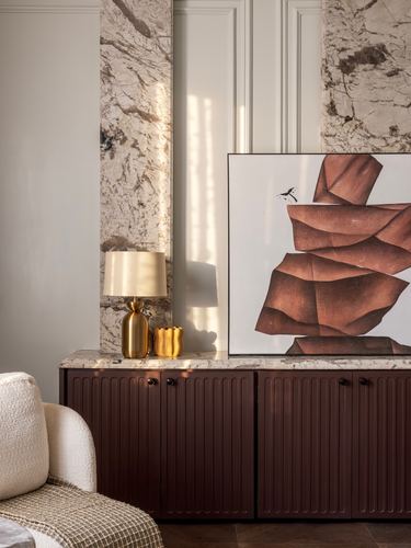 Marble-clad wall niche above a fluted maroon console with lamp and abstract art in sunlight