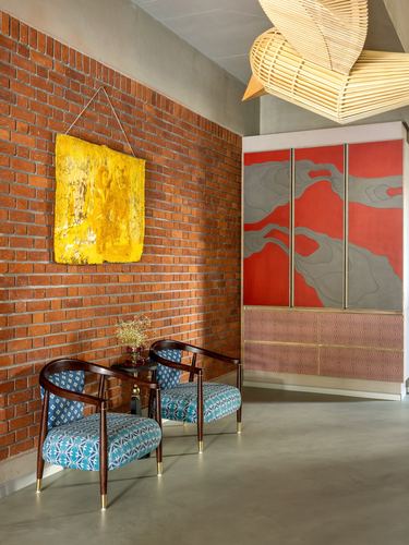 Brick-walled foyer with cane armchairs, yellow textile art, red painted cabinet and woven pendant light