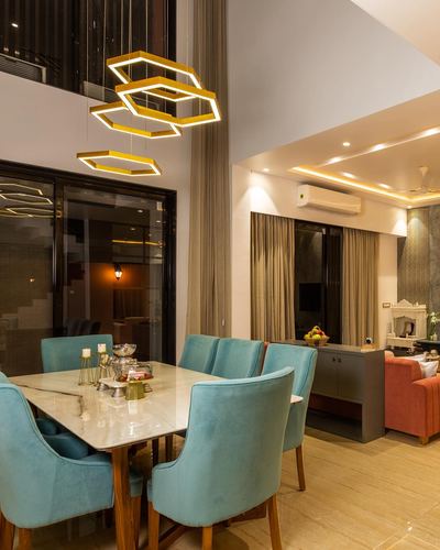 Dining area with teal velvet chairs, marble table, and geometric gold pendant lights under double-height ceiling
