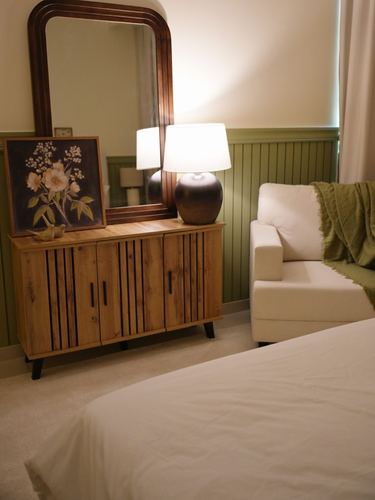 Bedroom corner with wooden sideboard, arched mirror, table lamp, floral art, and cream armchair against green wainscoting