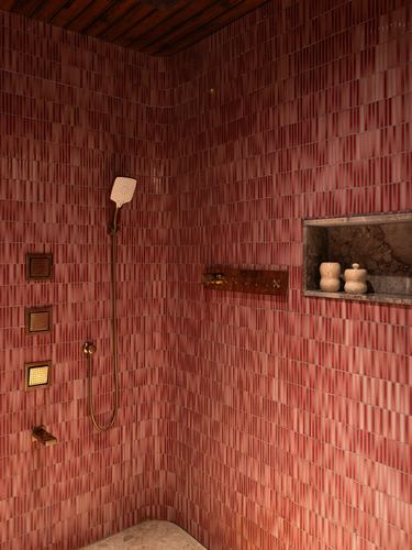 Corner of a shower stall clad in vertical pink ribbed tile with brass handheld shower and a stone niche holding small vessels