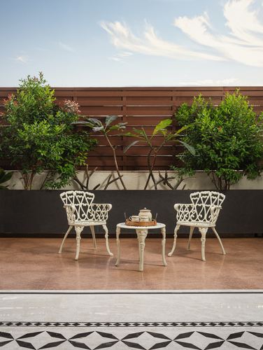 Two white wrought iron chairs and a round table on a terracotta terrace with plants and wooden slatted fence