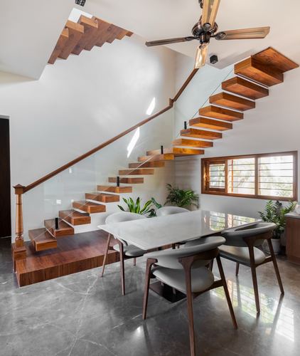Cantilevered wooden staircase with glass railing above a marble-top dining table in Bangalore villa