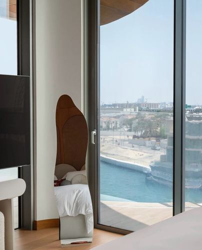 Sculptural wooden chair by floor-to-ceiling window overlooking Dubai waterfront from a neutral bedroom
