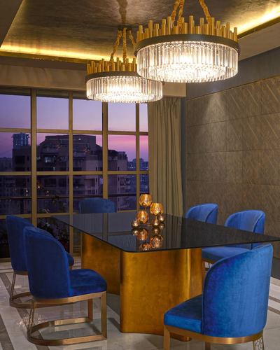 Blue velvet chairs around a glass and gold dining table under crystal chandeliers, dusk skyline behind