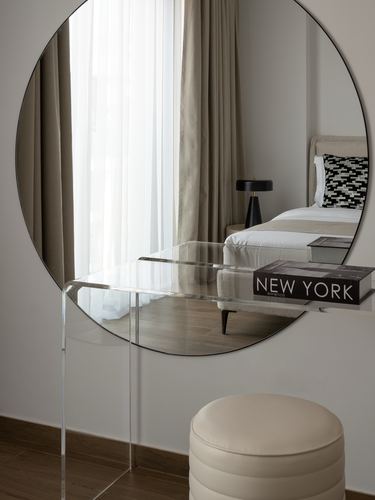 Round mirror above acrylic console reflecting a beige bed, with a leather pouf below in a neutral bedroom