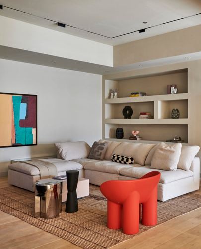 Beige living room with cream sectional, built-in shelving, and a red sculptural chair on jute rug