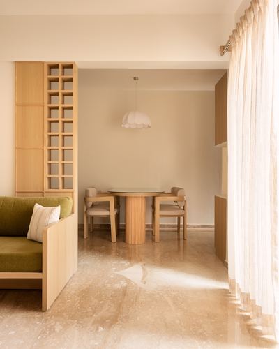 Sunlit dining nook with round wooden table, fluted base, scalloped pendant, olive sofa, marble floor