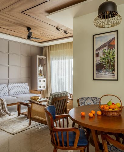 Living and dining area with wood-panelled ceiling, cane armchairs, round dining table with fruit basket, framed art beside pooja niche.