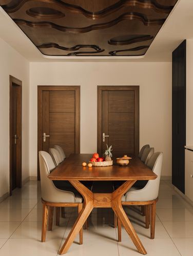 Dining table with cross-leg base under a carved wood-patterned false ceiling, flanked by two wooden doors.