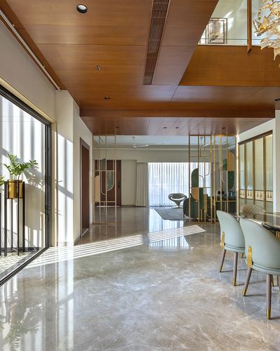 Double-height foyer with wood-clad ceiling, brass screen partition, marble-look floor and dining chairs under raking sunlight