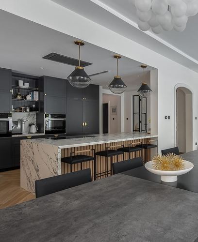 Black kitchen with waterfall marble island, brass pendant lights, and dark dining table in foreground