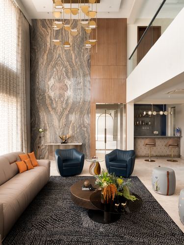 Double-height living room with bookmatched stone wall, brass pendant chandelier, and blue leather chairs