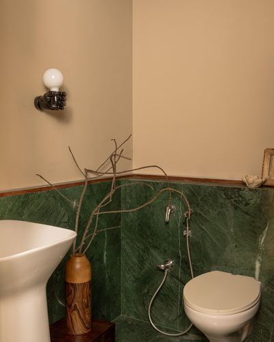 Bathroom corner with green stone dado, wall-hung toilet, hand-shaped sconce, and dry branches in wooden vase