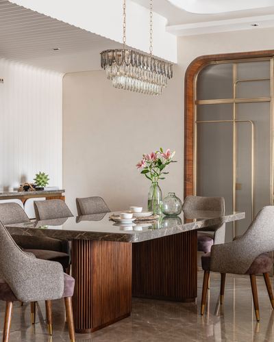 Dining table with fluted wood base and marble top under a crystal chandelier, arched brass-framed door beyond