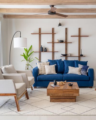 Living room with navy sofa, cream armchairs, wooden coffee table and asymmetric wall shelving under exposed beams