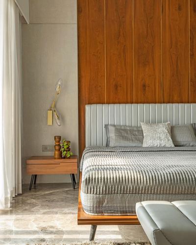 Bedroom with wood-panelled wall, grey channel-tufted headboard, wooden nightstand, brass sconce, marble floor.