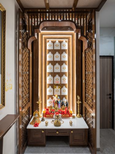 Wooden pooja unit with carved jali doors, backlit arched niche, and deity idols on marble ledge