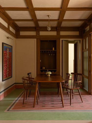 Wood-beamed dining room with windsor chairs, built-in bar niche, and a patterned rug on green flooring.