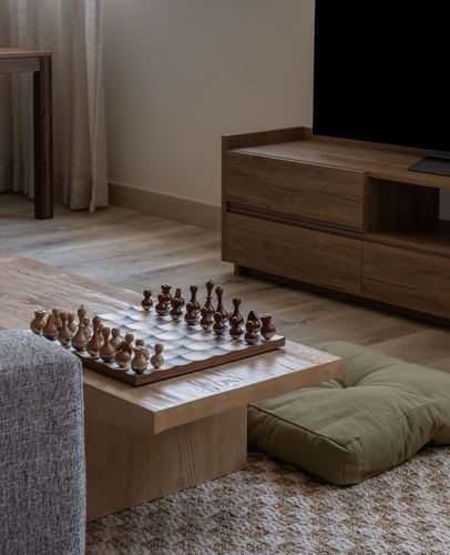 Low wood table with chess set beside floor cushion, wooden tv console in background