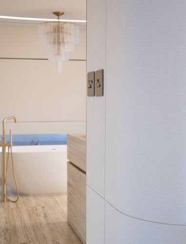 Curved linen-clad wall with switch plate framing a blurred bathroom with tub and chandelier beyond