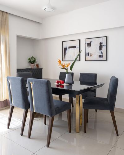 Dining room with black glass table on brass legs, six slate-blue velvet chairs, twin framed artworks on white wall.
