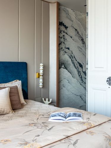 Bedroom corner with teal velvet headboard, floral linens, and a marble-clad wall glimpsed through an open door