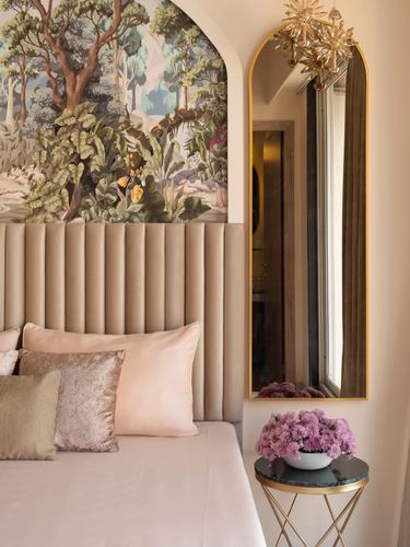 Blush channel-tufted headboard beneath an arched forest mural, gold mirror and marble side table beside it
