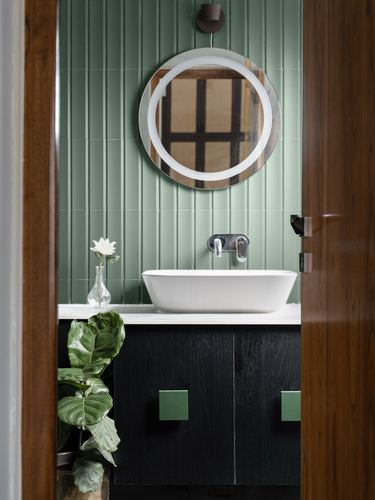 View through a wooden door into a powder room with green fluted tile, round mirror, and vessel sink over a dark green vanity.