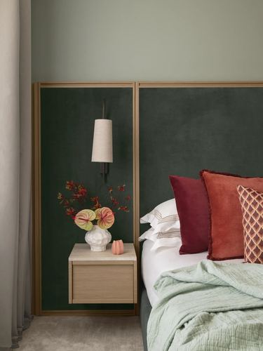 Green velvet headboard with wood trim, floating wood nightstand, sconce, and layered red pillows on a sage bed