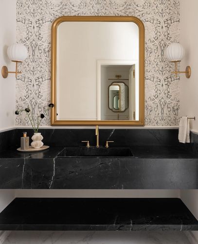 Bathroom vanity with black marble counter, gold arched mirror, and floral wallpaper